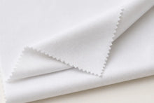 A close-up of folded white fabric with a fine ribbed texture and zigzag-cut edges, photographed on a light neutral background.