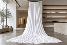 A large white fabric drape or curtain hanging from a wooden rod in a minimalist, modern textile showroom with concrete floors and shelving.