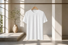 A plain white short-sleeve crewneck T-shirt hanging on a wooden hanger from a clothing rack, set in a minimalist, sunlit room with a window, plant, and concrete floor.