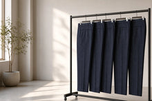 Four pairs of dark navy blue dress pants hanging on a black metal clothing rack in a bright, modern room with natural sunlight and a potted plant.