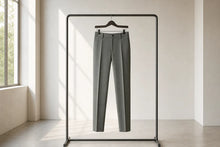 A pair of slim-fit gray trousers hanging on a black clothing rack in a minimalist, sunlit room with a window and light-colored walls.