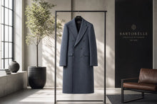 A long double-breasted coat in a muted blue-gray color with subtle vertical pinstripes, hanging on a minimalist black clothing rack inside a modern, sunlit interior. A brown leather chair is on the right, and a black wall panel displays the brand name 'SARTORELLE'.