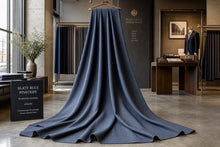 A slate blue ribbed knit fabric draped over a hanger in a boutique interior, with a sign reading 'SLATE BLUE PINSTRIPE' and a 'Heritage Wool Shop' wall display visible in the background.