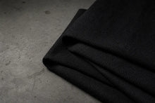 A close-up of folded black woven fabric, likely wool or a similar textured textile, laid on a dark gray concrete surface.
