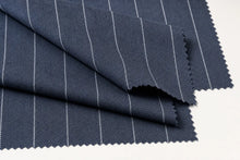 A close-up of folded navy blue fabric with a fine white herringbone weave and thin vertical white pinstripes, shown on a plain white background.