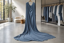 A draped slate blue fabric, likely a long dress or gown, hanging on a wooden hanger in a modern, sunlit boutique showroom. The fabric pools on the polished concrete floor, with racks of matching blue and white garments visible in the background.