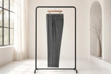 A pair of dark gray tailored trousers hanging on a minimalist black clothing rack in a bright, modern room with soft natural light.