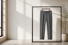 A pair of dark gray tailored trousers hanging on a black hanger inside a large black-framed mirror, set in a minimalist room with warm sunlight and shadows.