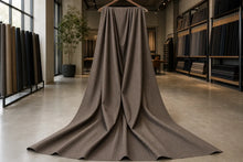 A large piece of taupe-brown fabric draped from a wooden hanger in the center of a fabric showroom, with shelves of other fabric rolls visible in the background.