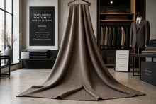 A draped taupe brown woven suiting fabric hangs from a wooden hanger in a modern, stylishly decorated clothing showroom. The fabric features a fine ribbed texture and is displayed prominently in the center, with suit racks, mannequins, and framed text boards visible in the background.