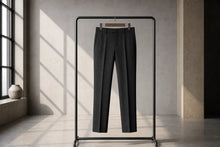 Dark charcoal gray trousers hanging on a minimalist black clothing rack in a modern, sunlit room with concrete walls and floor.