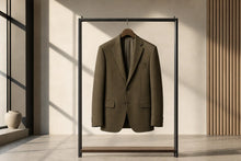 A tailored blazer in a muted olive-brown tone hanging on a minimalist black clothing rack inside a modern interior with warm sunlight and shadows.