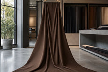 A large brown fabric drape hanging from a wooden hanger in a modern interior showroom with windows, shelving, and additional fabric swatches in the background.