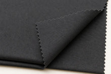 A close-up of folded black fabric with serrated edges, showing a fine twill texture, on a light neutral background.