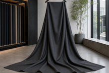 A large piece of dark charcoal gray fabric draped from a hanger in a modern, minimalist studio or showroom with large windows and a potted plant.