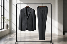 A dark gray two-piece suit, consisting of a single-breasted blazer and matching trousers, hanging on a black metal clothing rack in a bright, modern room with natural window light.