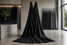 A long black fabric drape hanging from a hanger in a minimalist interior studio or showroom, with other dark fabric rolls visible in the background.