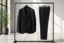A black two-piece suit, consisting of a single-breasted blazer and matching trousers, hanging on a minimalist black metal clothing rack in a bright, modern room with natural window light.