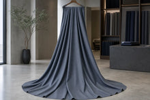 A long, floor-length gray fabric drape or curtain hanging from a wooden hanger in a modern interior showroom with shelves and a plant in the background.