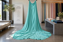 A long turquoise fabric dress form or draped garment displayed indoors in a fabric showroom, with a bench, plants, and hanging color swatches visible in the background.