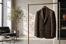 A brown plaid blazer jacket hanging on a gold metal clothing rack in a modern, sunlit room with a chair and potted plant in the background.
