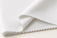 A close-up of folded white fabric with a fine woven texture and scalloped zigzag edges, photographed on a light neutral background.