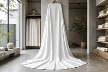 A long white fabric drape or curtain hanging from a wooden hanger in a minimalist, modern boutique interior with concrete floors, shelving, and large windows.