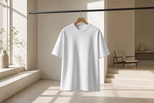 A plain white short-sleeve crewneck T-shirt hanging on a wooden hanger from a black clothing rack, set in a minimalist, sunlit beige room with a chair and window in the background.