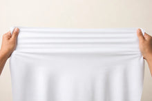 A pair of hands holding up a large, plain white sheet of fabric against a light beige background.