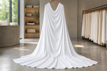 A white draped gown with a long train hanging on a wooden hanger in a boutique interior with clothing racks and shelves in the background.