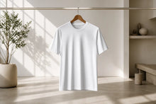 A plain white short-sleeve crewneck T-shirt hanging on a wooden hanger from a clothing rack, set in a minimalist, sunlit room with a potted plant and shelves in the background.