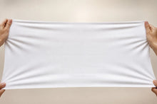 A plain white rectangular fabric sheet or towel is stretched horizontally by four hands against a light beige background.